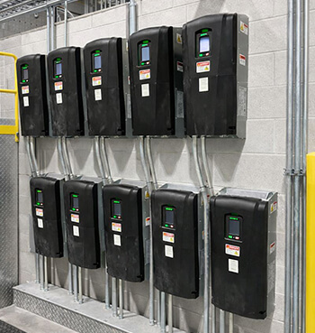 A row of ten black industrial variable frequency drives mounted on a concrete wall in a clean facility with organized conduit wiring.